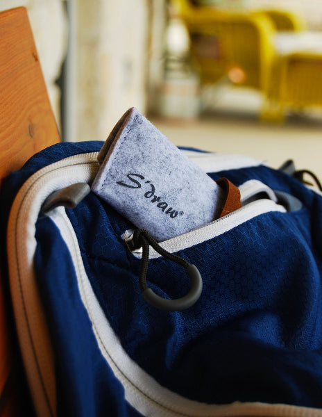 Image of a porch with a blue backpack with white zippers and in a pocket is a light heather grey wool travel case with the word S'draw printed on it