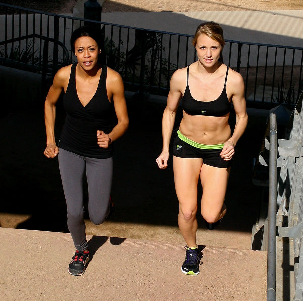 Image of two women running up steps, wearing a black tank top and grey leggings and a black sport bra and black running shorts with a bright green waistband folded over