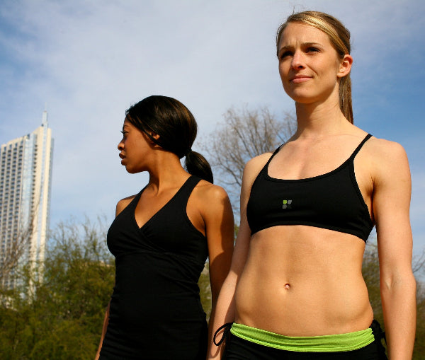 Image of two female models standing in a city park, wearing a black sport bra and black running shorts with a bright apple green waistband folded over and a black tank top