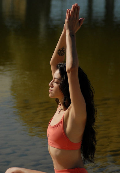 Side view image of a woman practicing yoga next to a lake, wearing coral orange color sport bra