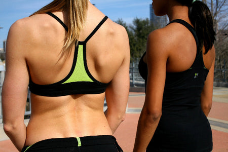 Back view of two female athletes standing on a running track, wearing a black sport bra with bright green mesh and black running shorts and a black racer back tank top